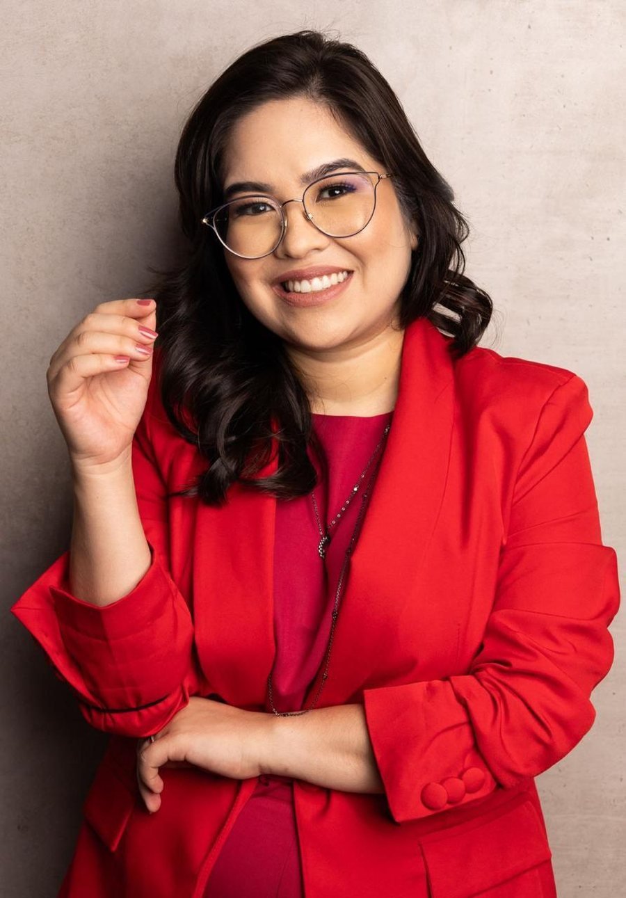 Dra. Paola Teruya sorrindo, de blazer vermelho e óculos, com os braços cruzados